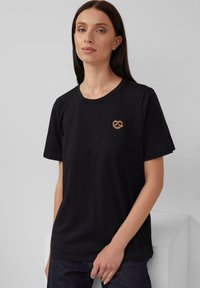 Zwarte katoenen T-shirt met korte mouwen, met een gouden, verfijnd ontworpen pretzel-embleem op de linkerborst.