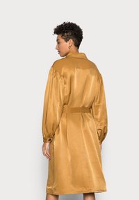 Vestido de satin amarelo mostarda com mangas longas e bufantes, uma gola e um cinto na cintura. Apresenta pregas nas costas e punhos com botões.