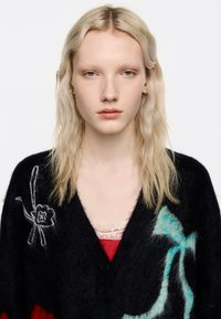 Czarny kardigan wełniany z wielokolorowymi kwiatowymi i wstążkowymi wzorami. Miękka faktura, zapinany na guziki z szerokimi klapami. Na warstwie znajduje się czerwona bluzka.