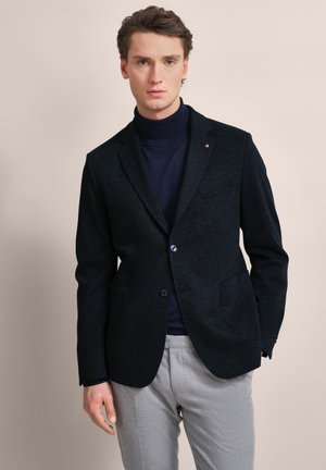 Marineblazer mit Struktur, zwei vorderen Taschen, Ein-Knopf-Verschluss und Innenfutter, kombiniert mit einem dunklen Rollkragenpullover und grauen Hosen.