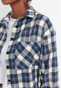 Modrá a bílá kostkovaná flanelová košile s černými detaily, s knoflíky, límečkem a přední kapsou. Měkká textura, pohodlný střih.