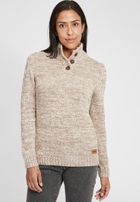 Beige strikket genser med høy hals, teksturert mønster, tre brune knapper på kragen, og et lite merke på kanten.