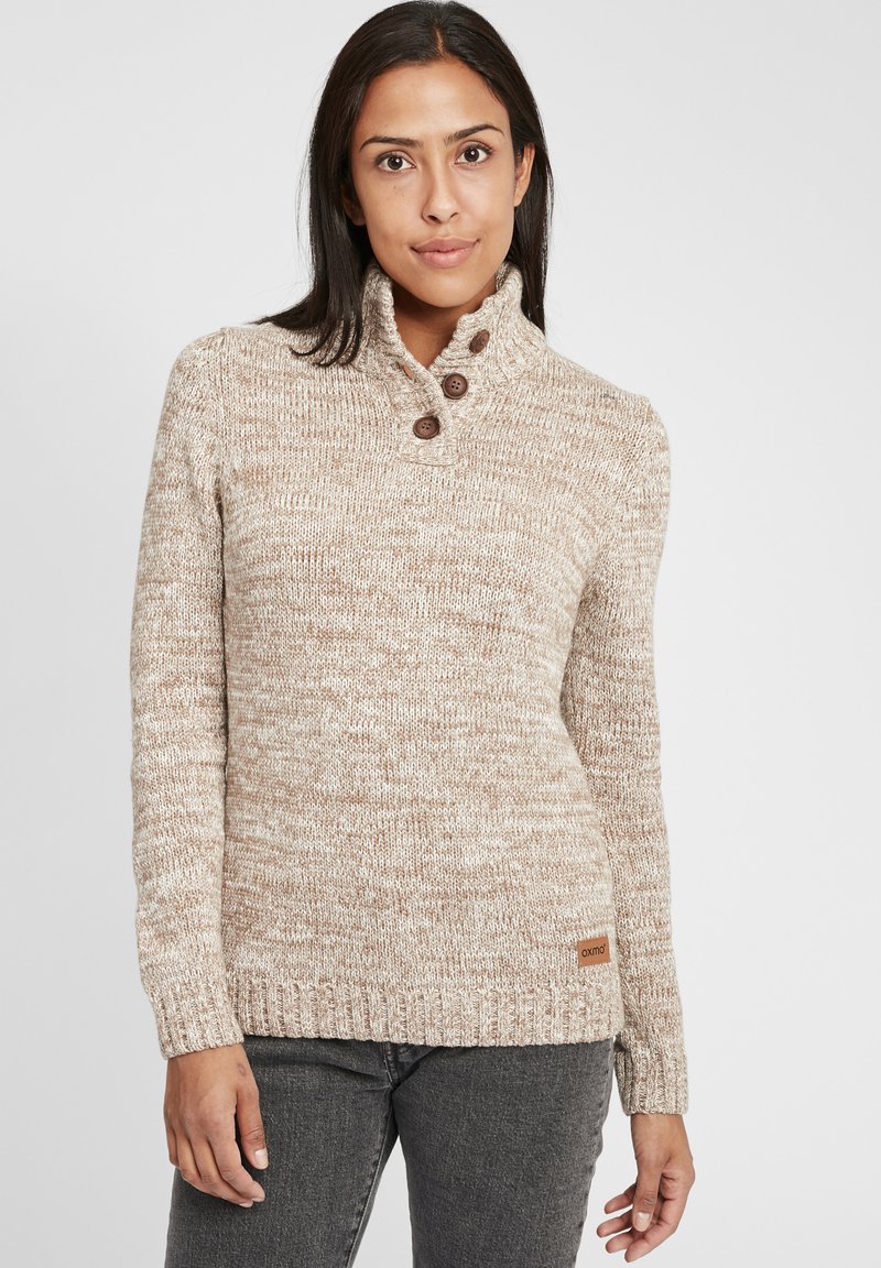 Beige strikket genser med høy hals, teksturert mønster, tre brune knapper på kragen, og et lite merke på kanten.