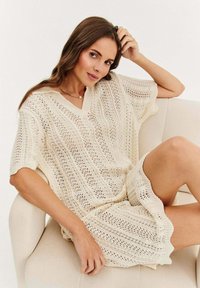 Sukienka dzianinowa z dekoltem w serek, krótkimi rękawami oraz ażurowym wzorem w kolorze kremowym, z falbaniastym wykończeniem i luźnym krojem.