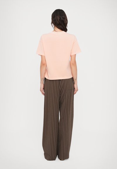 Ανοιχτό ροδακινί κοντομάνικη μπλούζα συνδυασμένη με καφέ ριγέ παντελόνες φαρδιάς γραμμής. Η μπλούζα έχει χαλαρή εφαρμογή και στρογγυλή λαιμόκοψη.