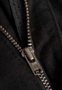 Tela negra con un zipper metálico con un tirador YKK, costuras visibles a lo largo de los bordes, mostrando una apariencia texturizada.