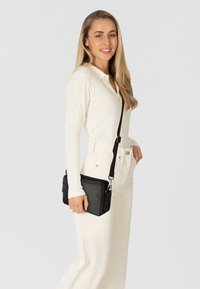 Borsa a tracolla nera sintetica con superficie testurizzata, cinturino regolabile e accessori in argento, tenuta da una persona che indossa una maglietta bianca a coste e pantaloni chiari.