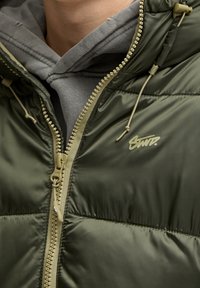 Chaqueta puffy verde oliva con cremallera al frente, detalles en color beige y un logotipo. Superficie texturizada con capucha ajustable y una sudadera gris por debajo.