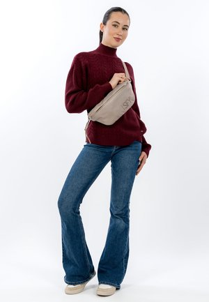 Beige strukturierte Umhängetasche mit Reißverschluss, Tragegurt und Logodetail. Das Modell trägt einen kastanienbraunen Pullover und ausgestellte Jeans.