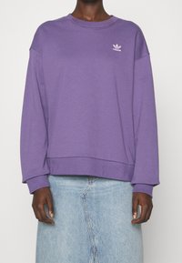 Fialová mikina s kulatým výstřihem a dlouhými rukávy, na levé části hrudi s malým bílým logem Adidas. Materiál vypadá měkký a strukturovaný.