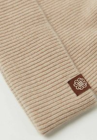 Beige ribbad stickad tyg med en mjuk textur och en liten brun etikett med en vit logotyp.