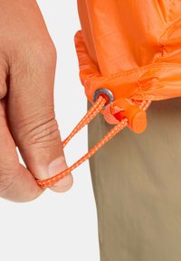 Orange lättviktsjacka med dragsko och snörning. En hand håller i den flätade dragskon som har en rostfri ögla.