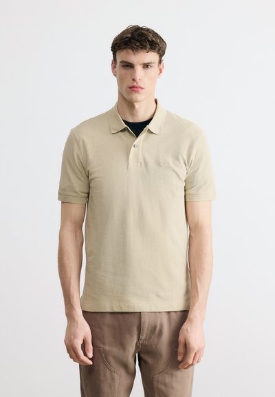 Jeune homme portant un polo beige à manches courtes et un pantalon marron, debout devant un fond clair uni, faisant face à l'avant.