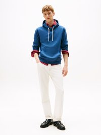 Sudadera azul con cordones blancos, sobre una camisa de cuadro roja. Combinada con pantalones blancos y zapatos negros, de pie contra un fondo blanco.