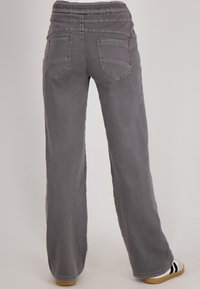 Achteraanzicht van een persoon die losse grijze broeken draagt met gestikte achterzakken en witte sneakers met zwarte strepen.