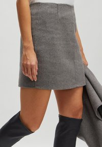 Grauer Wollrock mit taillierter Passform, knielangem Saum und dezenten Nähten. Begleitet von einem dunkelgrauen Mantel und kniehohen schwarzen Stiefeln.