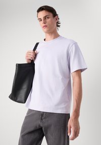 T-shirt en coton lavande à manches courtes, avec un logo sur le devant, associé à un pantalon en denim gris et un sac fourre-tout en cuir noir.