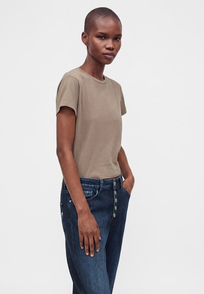 Femme aux cheveux courts portant un t-shirt taupe uni et un jean bleu foncé taille haute avec des boutons, debout avec une main dans la poche.