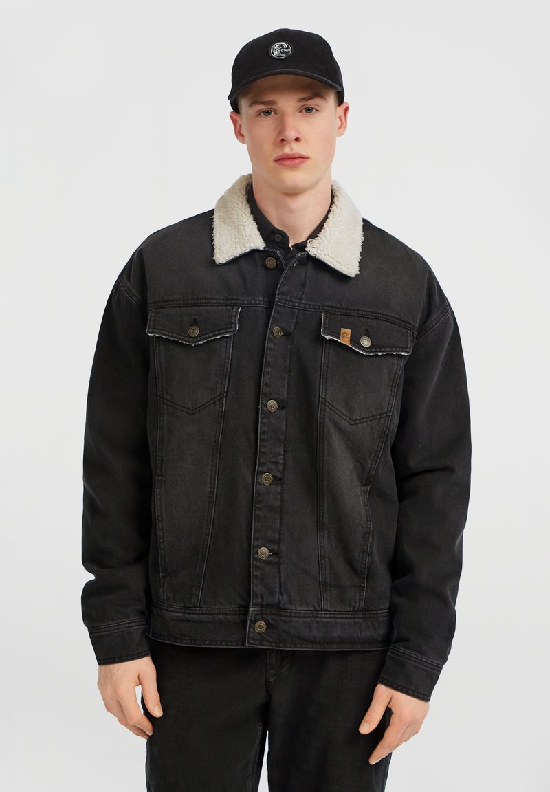 Ung man med en svart denimjacka med vit fleecekrage och en svart keps, står mot en enkel ljusgrå bakgrund.