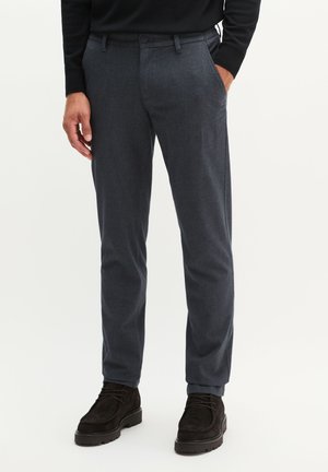 Mann trägt dunkelblaue strukturierte Hose mit schwarzen Schnürstiefeln und einem schwarzen Langarmoberteil, steht mit einer Hand in der Tasche.
