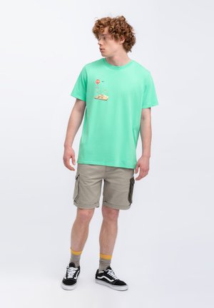 T-shirt in cotone verde menta con una piccola stampa grafica sul petto, abbinata a pantaloni cargo color khaki fino al ginocchio e scarpe da ginnastica nere.