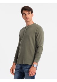 Olivgrünes Langarm-Henley-Shirt mit drei Knöpfen, rundem Ausschnitt und weichem Stoff, kombiniert mit blauen Jeans und einer Uhr.