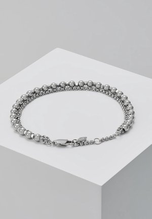 Bracelet ton argent avec deux chaînes, une perlé et une à maillons, avec un fermoir mousqueton et une petite étiquette gravée sur une surface blanche.