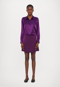 Lilla satin skjorte med knapper og lange ærmer, parret med en matchende lilla mini-skjørt. Glat tekstur, struktureret pasform og minimal detaljering.