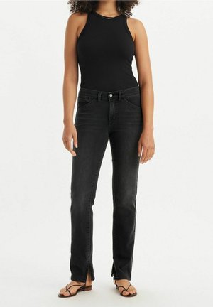 Femme portant un haut noir sans manches, un jean noir taille haute avec des fentes aux chevilles à l'avant et des sandales noires à brides, debout sur fond blanc.