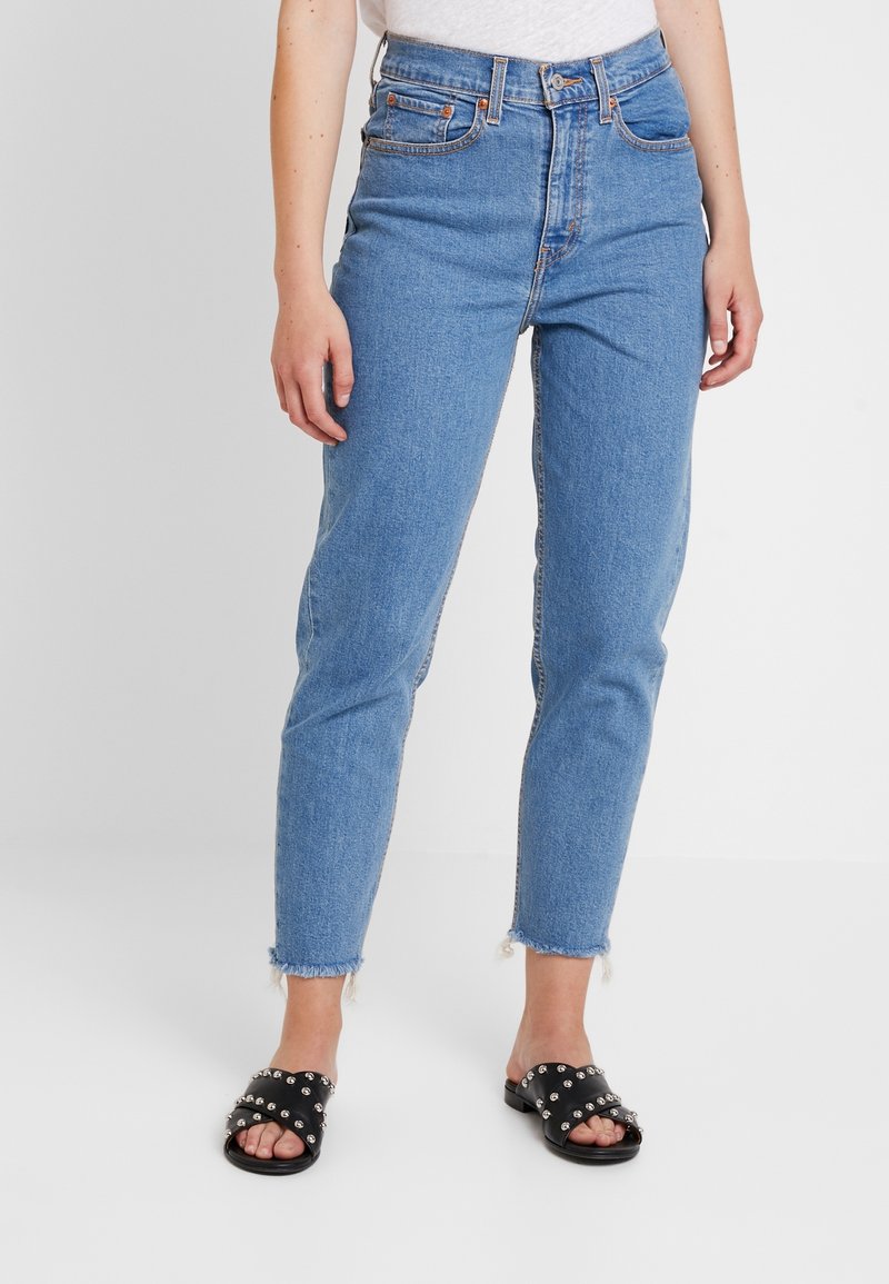 Vaqueros azules de mezclilla con cintura alta, pierna recta y dobladillo deshilachado. Combinados con sandalias negras con tachuelas plateadas.