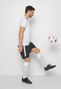 Baltas trumpas sportinis marškinėliai su juodais akcentais, juodos šortai su balta šonine juostele, balti iki kelių siekiantys kojinės ir juodi futbolo batai.