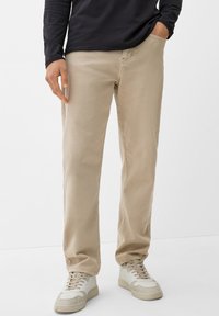 Pantalones de sarga de algodón beige con un corte relajado, que presentan costuras de doble puntada y bolsillos laterales, combinados con zapatillas deportivas blancas.
