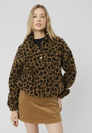 Pull à motifs léopard brun avec un col haut, fabriqué en tissu texturé. Associé à une jupe en velours côtelé beige. Trois boutons au niveau du col.