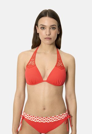 Mujer con bikini rojo, top de crochet con cuello halter y braguitas con lazos laterales y cinturilla estampada, de pie frente a un fondo claro y liso.