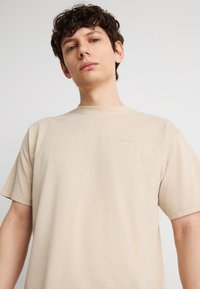 Νέος άνδρας με κοντά σγουρά μαλλιά, που φοράει μια απλή μπεζ μπλούζα Levi's, στέκεται μπροστά σε ουδέτερο φωτεινό φόντο.