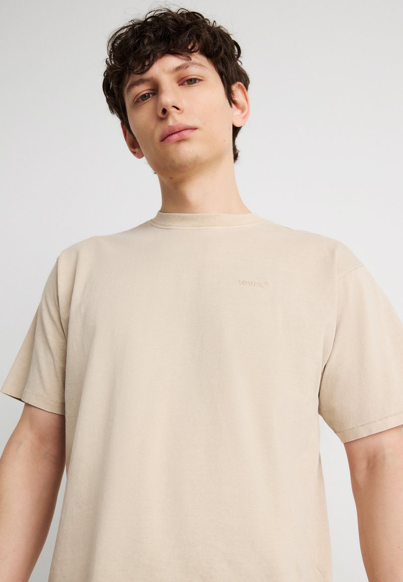 Νέος άνδρας με κοντά σγουρά μαλλιά, που φοράει μια απλή μπεζ μπλούζα Levi's, στέκεται μπροστά σε ουδέτερο φωτεινό φόντο.