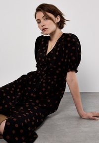 Zwarte jurk met bruine stippen, knoopsluiting aan de voorkant, korte pofmouwen en een getailleerde taille. Gladde stof met een relaxte pasvorm.