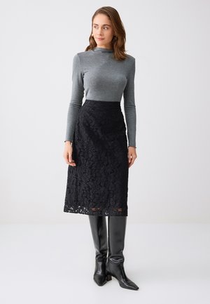 Femme portant un haut moulant gris à manches longues, une jupe midi en dentelle noire et des bottes en cuir noires montant jusqu'aux genoux, debout devant un fond uni.