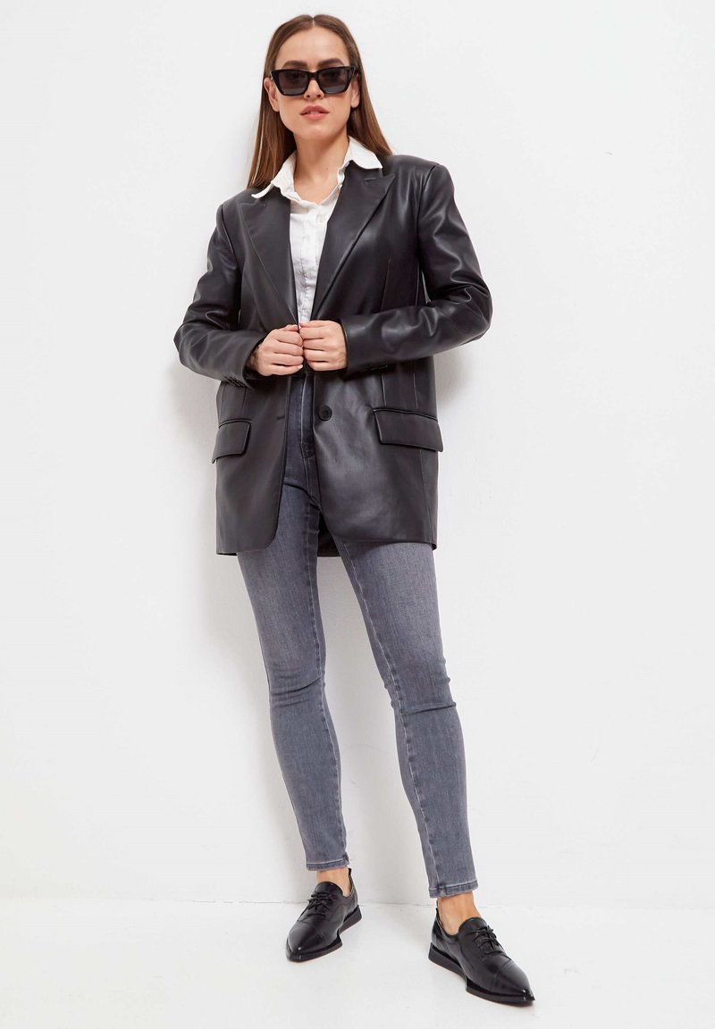 Blazer de cuero negro con un solo botón y dos bolsillos frontales, combinado con jeans ajustados grises y zapatos de cordones negros.