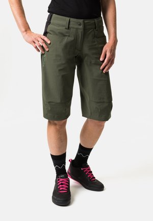 Olijfgroene shorts met een recht model, voorzien van voorzakken en een zwarte tailleband. Gecombineerd met zwarte schoenen en roze veters.