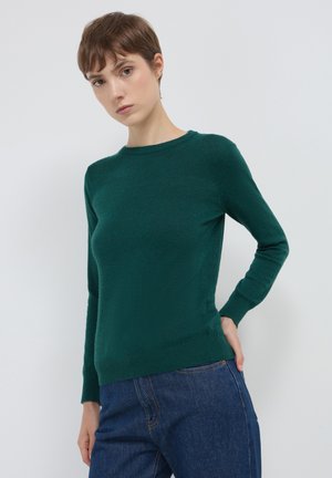 Giovane con i capelli corti castani che indossa un maglione aderente a maniche lunghe di colore verde scuro e jeans blu, in piedi contro uno sfondo bianco uniforme.