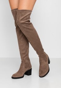 Žena v taupe semišových over-the-knee botách s blokovými podpatky, stojící na jednobarevném bílém pozadí.