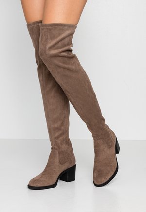 Žena v taupe semišových over-the-knee botách s blokovými podpatky, stojící na jednobarevném bílém pozadí.