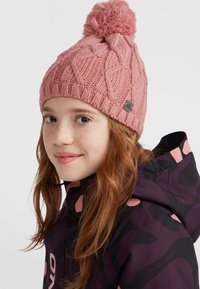 Rosa stickad mössa med ett geometriskt mönster, utrustad med en fluffig pom-pom på toppen och en liten metallisk logotypetikett på sidan.