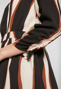 Schwarz, creme und rostfarben gestreifter Stoff mit texturierter Oberfläche; verfügt über einen gerafften elastischen Ärmel und einen passenden Gürtel in der Taille.