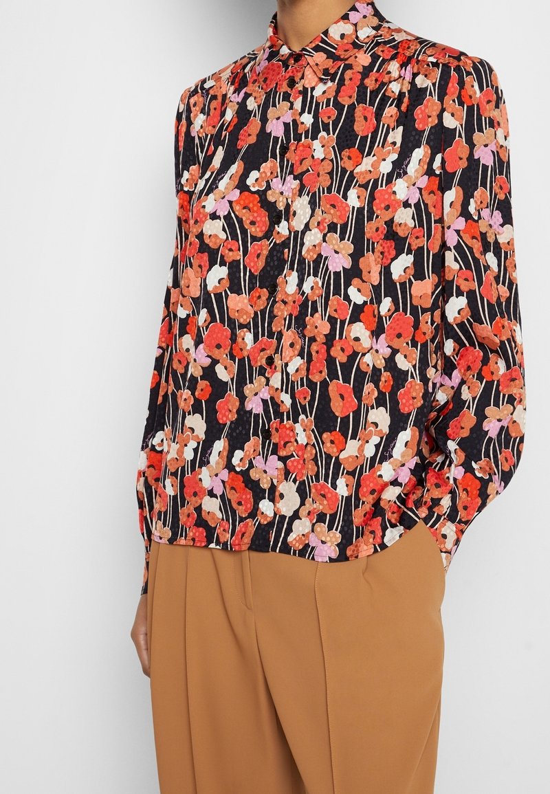 Blouse à manches longues fleurie avec des fleurs rouges, orange, roses et blanches sur fond noir, associée à un pantalon camel taille haute.