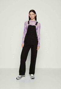 Kvinde står iført sorte overalls, en lilla langærmet bluse og sølvsnøresko foran en ensfarvet hvid baggrund.