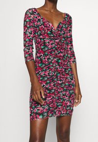 Robe courte fleurie à manches longues, présentant un fond noir, des motifs de feuilles roses et vertes, avec des fronces sur le devant.