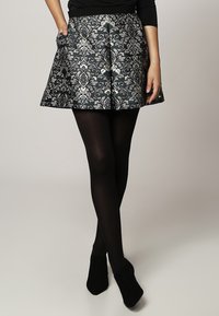 Femme portant des collants noirs, des chaussures noires et une jupe courte à motifs noir et argent, avec les mains partiellement dans les poches.