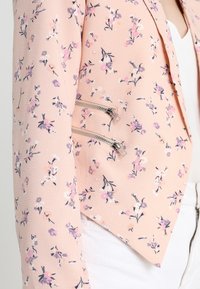 Světle růžový květinový blazer s malými fialovými a bílými květinami, se dvěma vodorovnými stříbrnými zipy poblíž pasu, nošený přes bílý top.